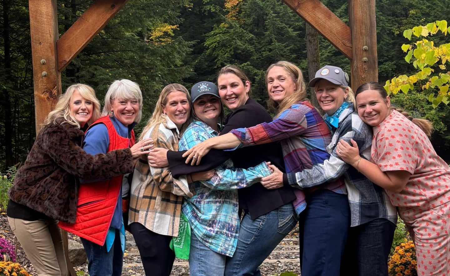 A group of friends hugging and smiling together under a wooden arch in the woods